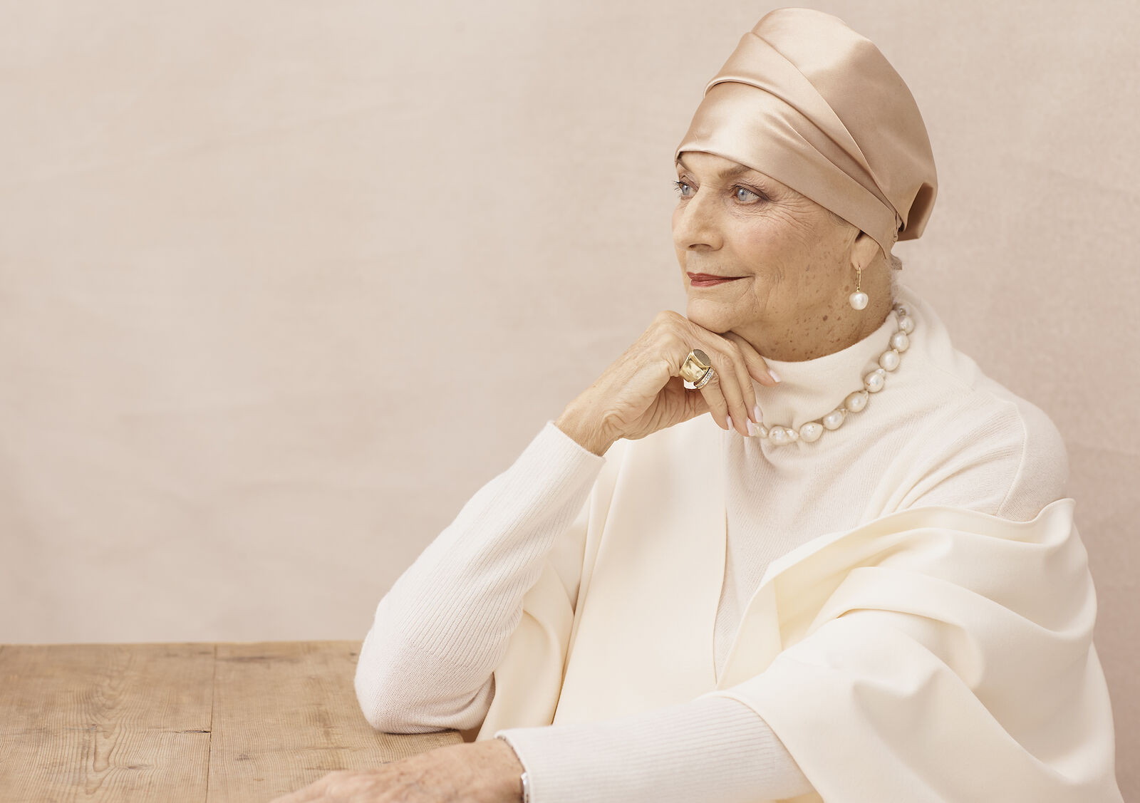 Elegant senior woman in a cream-colored outfit, headscarf, and pearl necklace, posing indoors with hand on her chin.