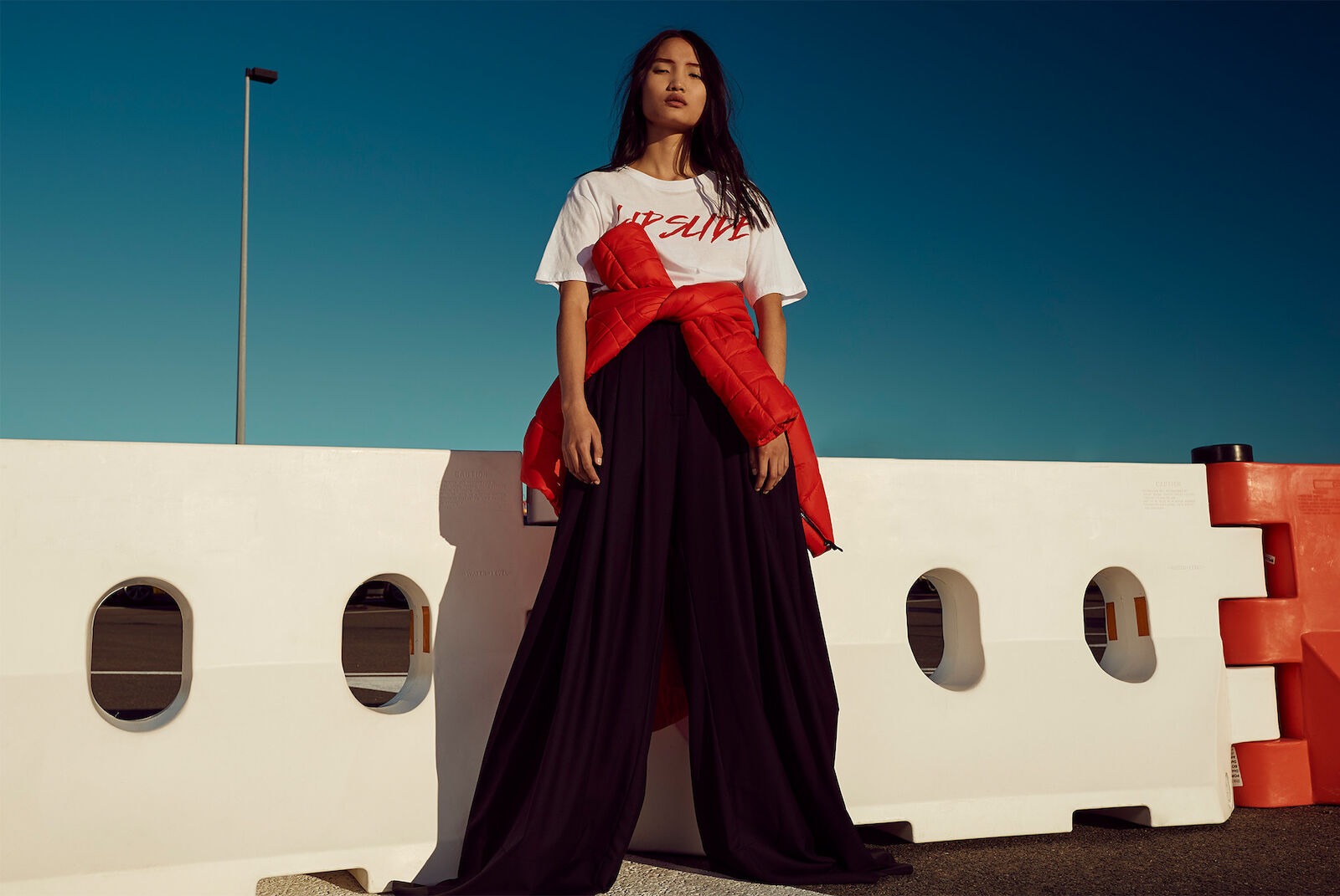 Woman in wide-leg pants and t-shirt poses in front of a barrier with blue sky. Red jacket tied around her waist adds a pop of color.