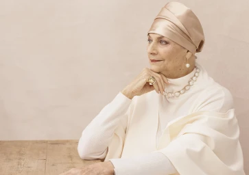 Elderly woman in a satin headwrap and white turtleneck posing with pearl jewelry against a neutral backdrop
