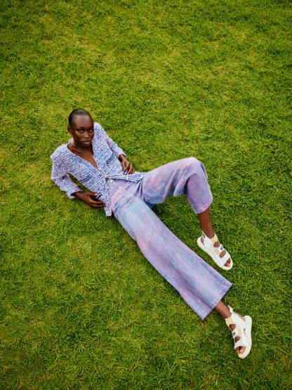 A dark-skinned woman lounges on a vibrant green lawn, wearing patterned shirt and wide pants, complemented by chunky white sandals.