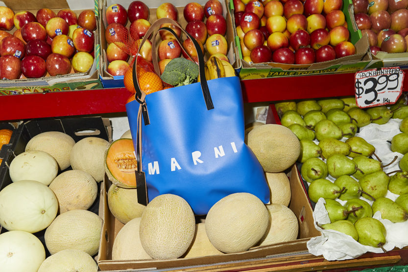 Close-up of a blue Marni shopping bag filled with fruits and vegetables, placed amidst cantaloupes at a market, creating a still life.