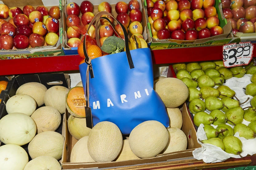 Close-up of a blue Marni shopping bag filled with fruits and vegetables, placed amidst cantaloupes at a market, creating a still life.