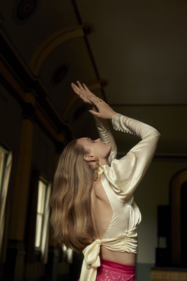 A woman in a white top and pink skirt raises her hands indoors, under dim lighting. Her long blonde hair is wavy as she looks up.
