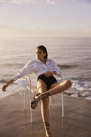 Woman poses on chair in black shorts and white top with ocean backdrop, lace-up sandals, and wet hair with natural lighting.
