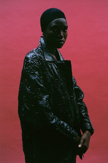 A person with dark skin and a black hat poses in front of a pink background, wearing a black floral embossed coat.