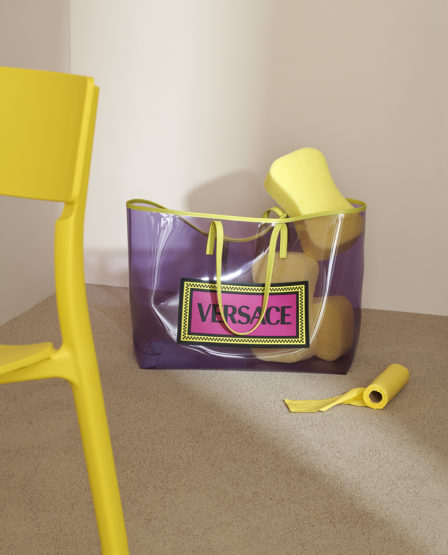 Versace purple tote bag filled with yellow sponges next to a yellow chair and a roll of yellow tape against a beige backdrop.
