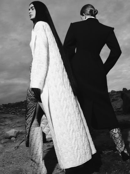 Monochrome fashion shot of two women outdoors. One in a white textured coat with long dark hair, the other in a black coat, back to camera.