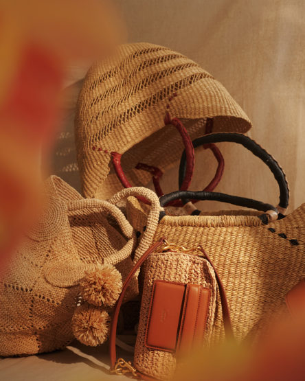 Still life shot of straw tote bags with leather handles and embellishments in warm neutral tones against a light background.
