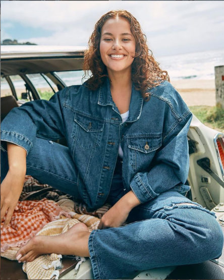 Woman in denim jacket and jeans poses in back of vehicle with sandy beach and ocean in background on a sunny day.