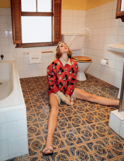 Woman sitting cross-legged on a patterned floor in a bathroom, wearing a red Mickey Mouse sweater and striped shirt with water stream.