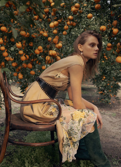 Woman in dress with floral detail sitting on wooden chair in orange grove wearing studded belt and green suede boots.