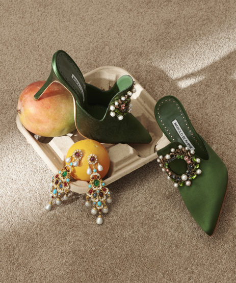 Elegant green high-heeled mules with bejeweled embellishments sit on an egg crate with fresh mango and mandarin oranges and drop earrings.