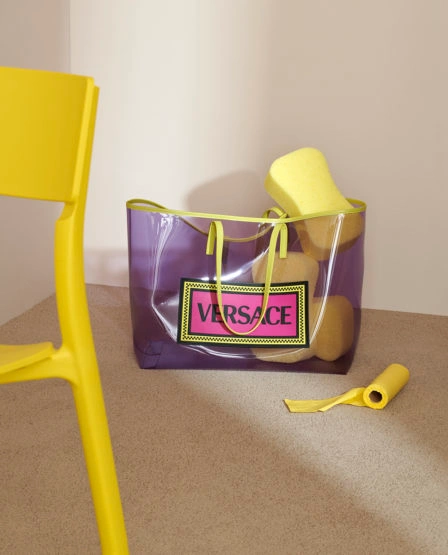 Transparent purple Versace tote bag filled with yellow sponges resting on a beige carpet against a plain wall.