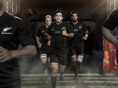 Rugby players in black uniforms run onto the field from a dark tunnel with fog. 'All Blacks' and 'AIG' logos are visible.