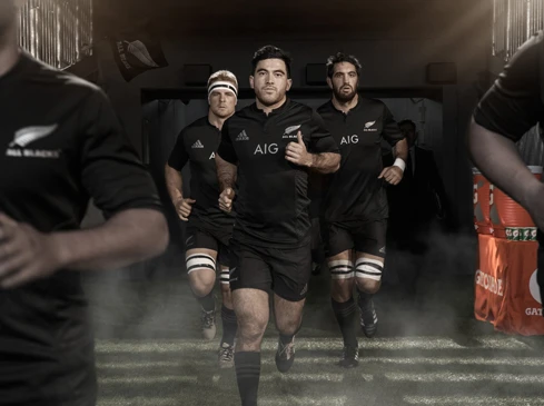Rugby players in black uniforms run onto the field from a dark tunnel with fog. 'All Blacks' and 'AIG' logos are visible.
