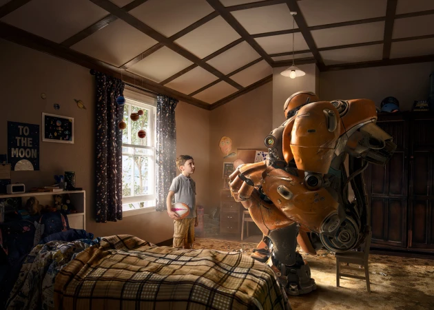 Young boy gazes at an orange robot crouched in his bedroom, holding a chair. Room is bathed in warm light with planets on the wall.