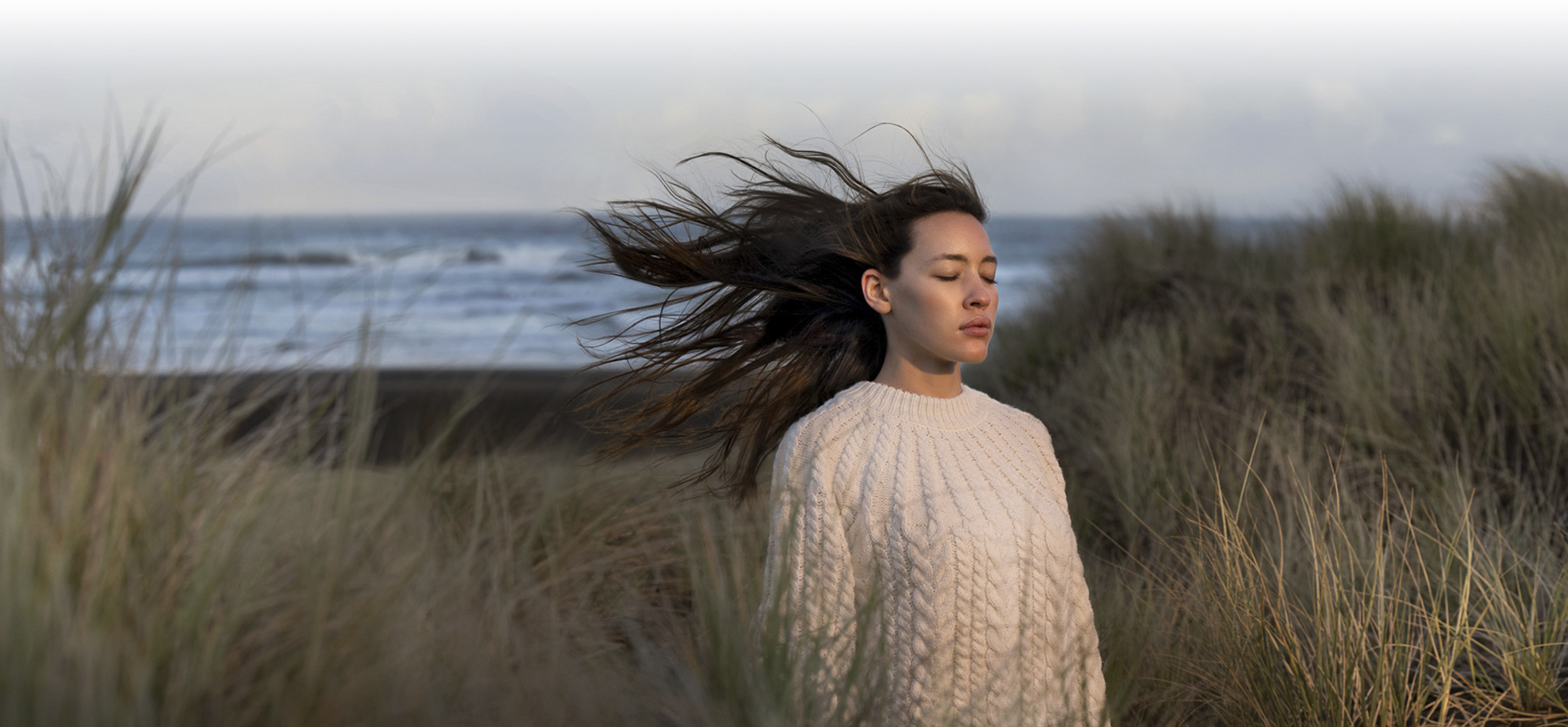Woman in white cable-knit sweater standing amongst dune grass at the beach with eyes closed in a portrait style photo.