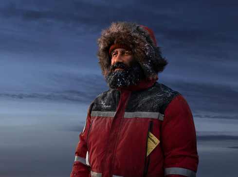 Man in a fur hat and winter coat stands outside against a dark sky, dusted with snow or frost on his face and clothing.