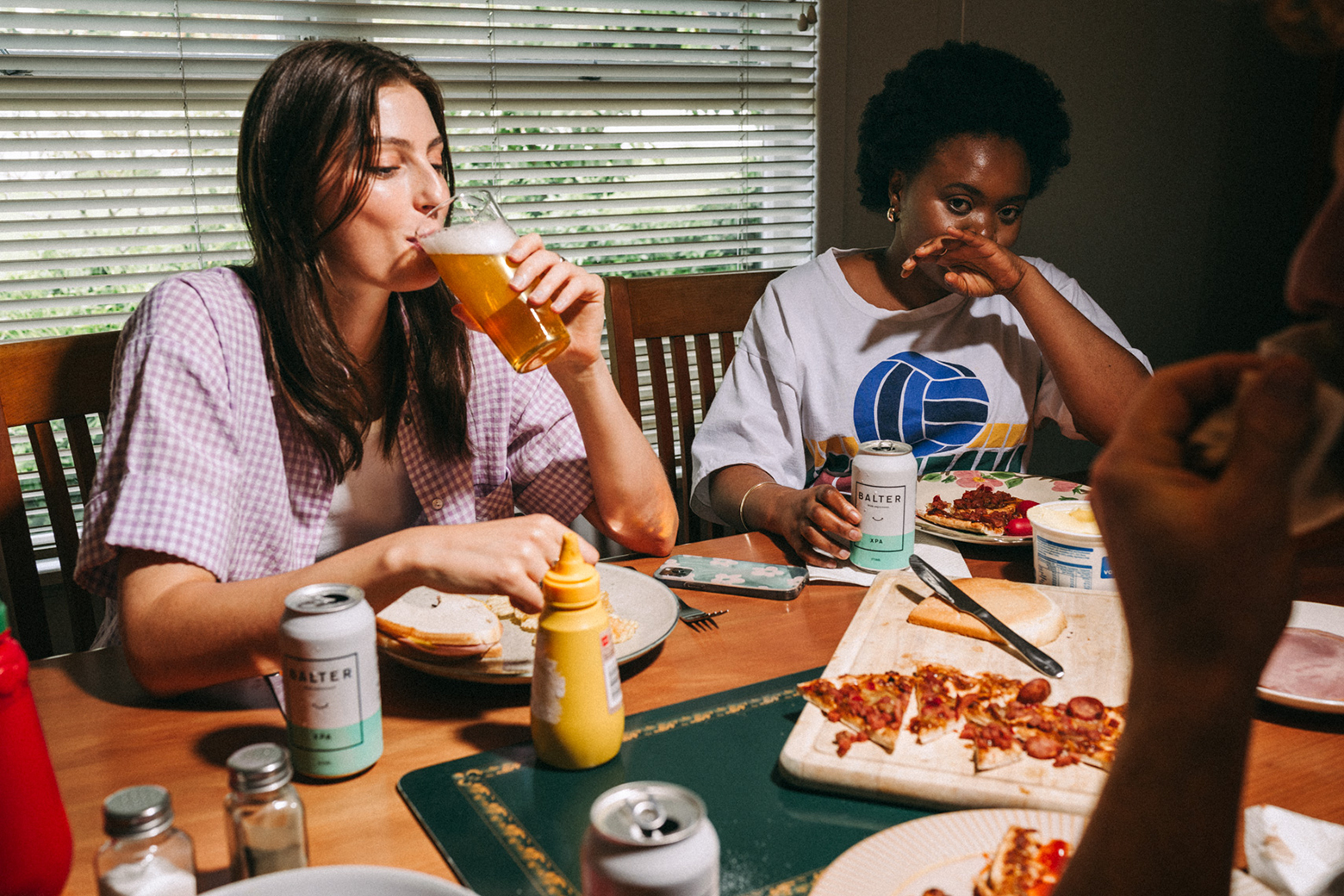 Two women and an unidentifiable person share a table with food, drinks, and cans of Salter beverages at an indoor gathering.