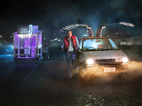 A man in an orange vest running toward a modified car with gull-wing doors, with a neon pay station glowing in the background.