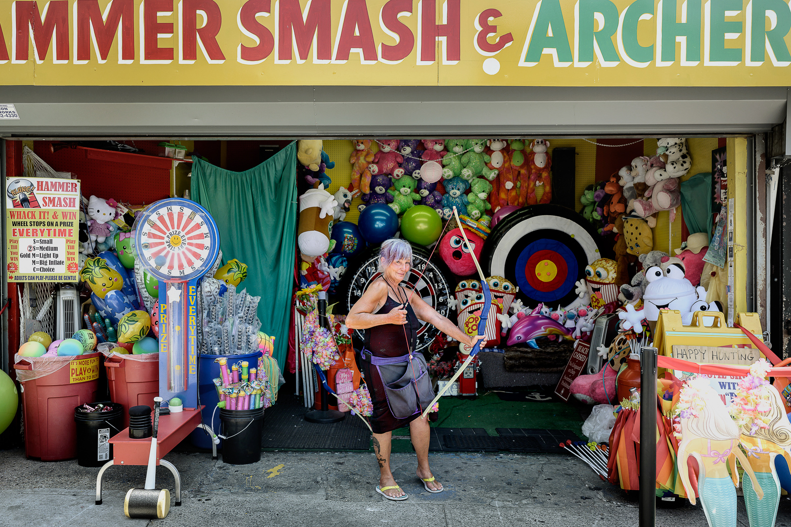 Elderly woman with bow and arrow at an amusement park game stall filled with stuffed animals and balloons, shot in an eye-level outdoor setting.
