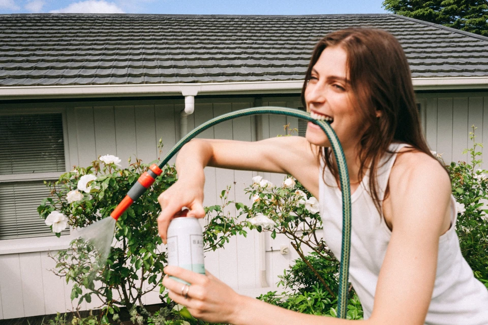 A woman stands outdoors, holding a watering can and a garden hose in her mouth, surrounded by greenery, with a house in the background.