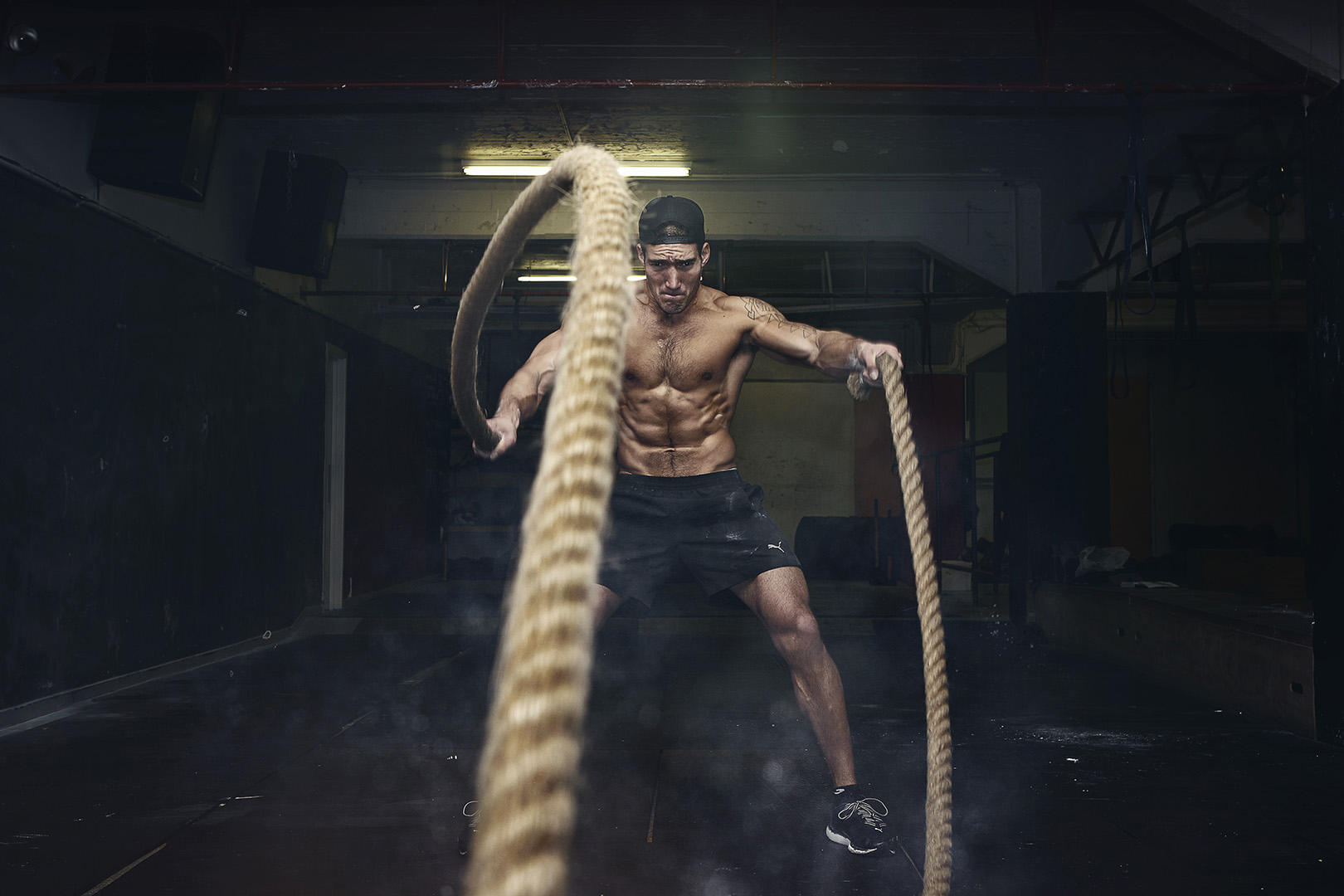 Muscular man does a battle rope workout in a dark, moody gym with dynamic lighting and visible abs, highlighting fitness and determination.