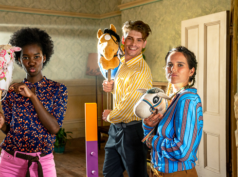 Three adults playfully pose indoors with toy stick horses, wearing colorful shirts and pink pants, in a light-filled room with minimal furniture.
