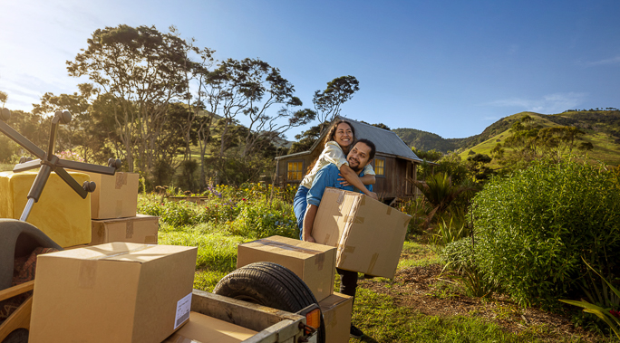 A couple moving to a new home in a rural area, hugging and carrying a cardboard box in front of a house and trailer full of furniture.