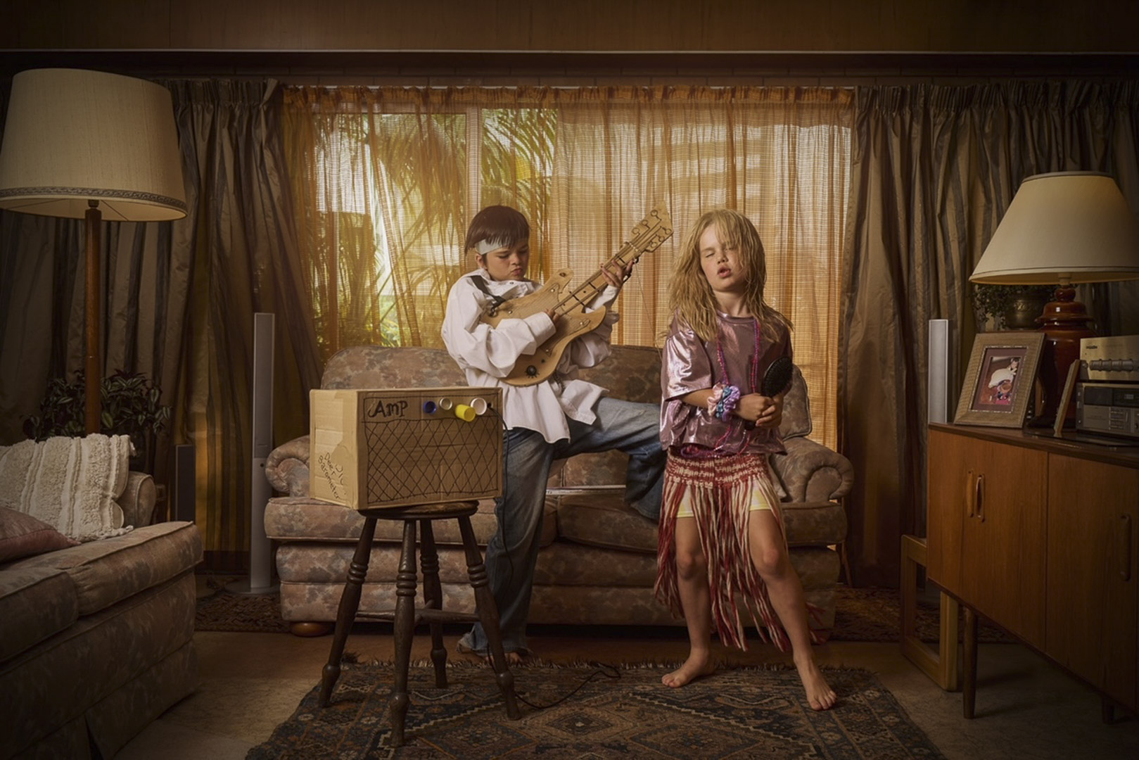 A boy plays a cardboard guitar and a girl sings into a hairbrush in a retro-styled living room with vintage furniture and curtains.