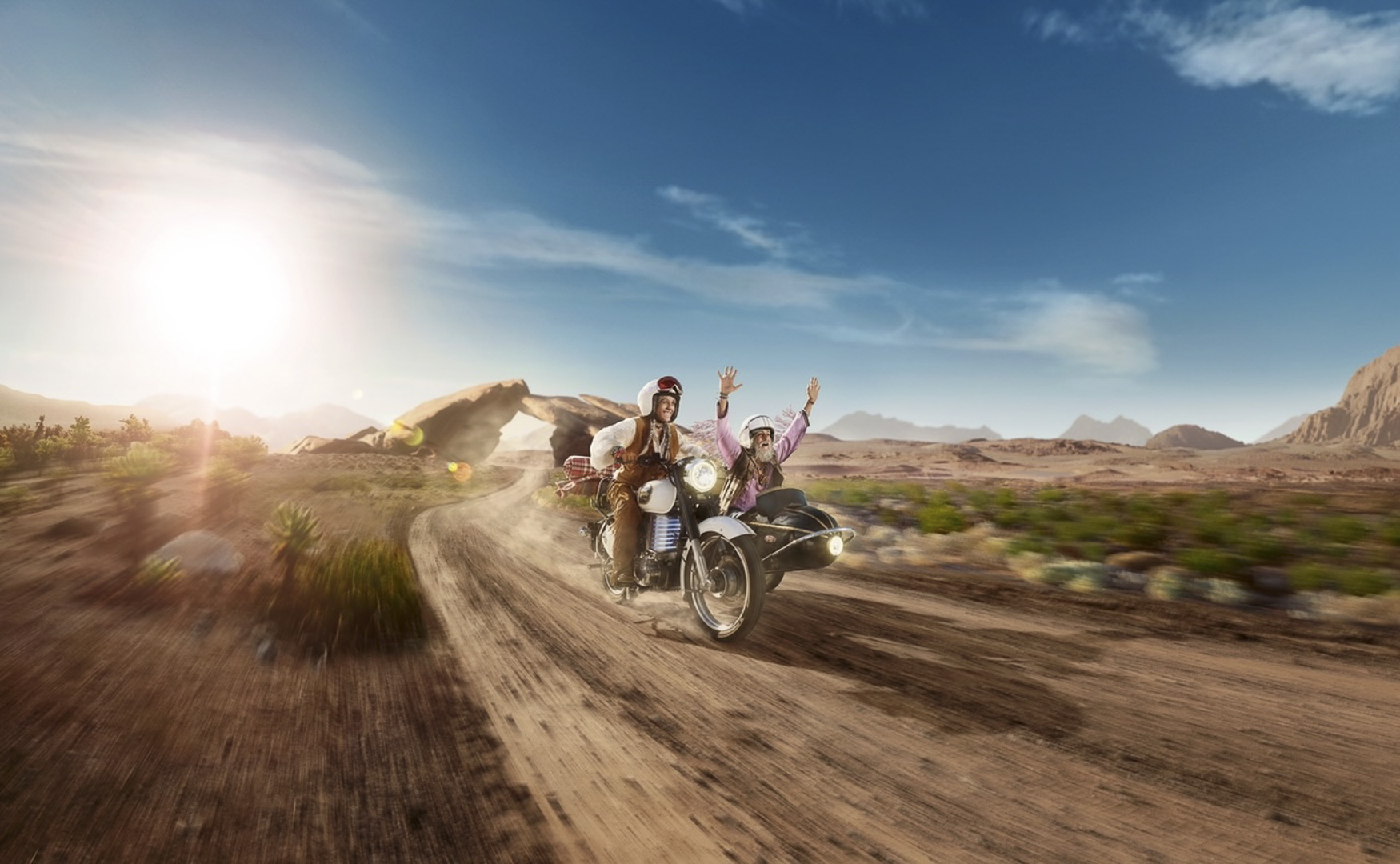 Two people ride a motorcycle with a sidecar on a dirt road in a desert landscape under a blue sky, one with raised arms.