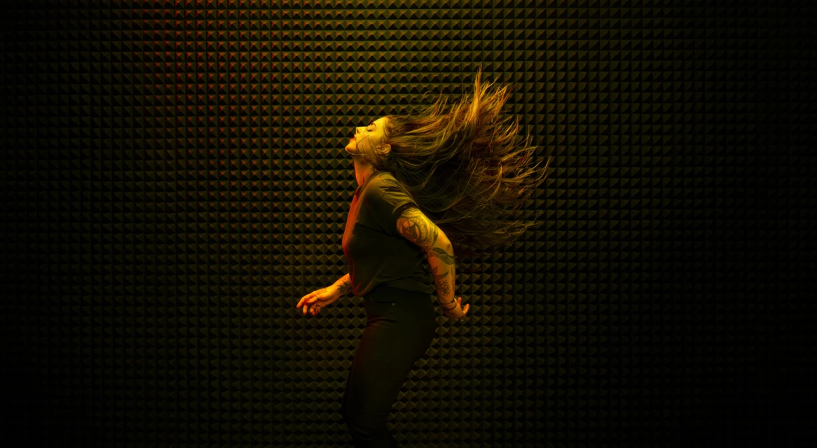 Adult female with tattoos tosses long brown hair in a dark room illuminated by yellow and orange light against a textured background.