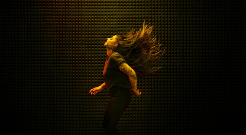 Adult female with tattoos tosses long brown hair in a dark room illuminated by yellow and orange light against a textured background.