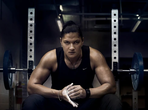 Muscular woman sits in a gym with barbell equipment in the background, wearing a black tank top and black bottoms, hands coated in chalk.