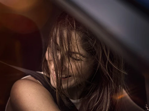 Woman sitting in a car with her hair covering her face, wearing a seatbelt, interior shot with blurred background, nighttime.
