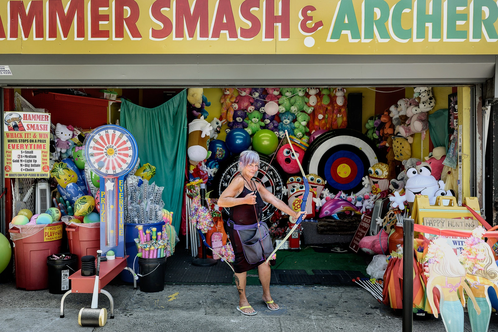 Elderly woman holding a blue bow in front of a carnival game booth filled with plush prizes and targets.