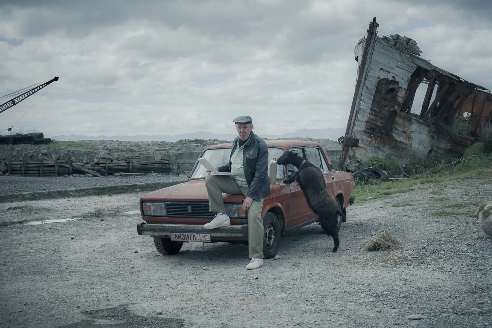 Elderly man seated on a red car with a black goat, near a ruined wooden shipwreck and crane.