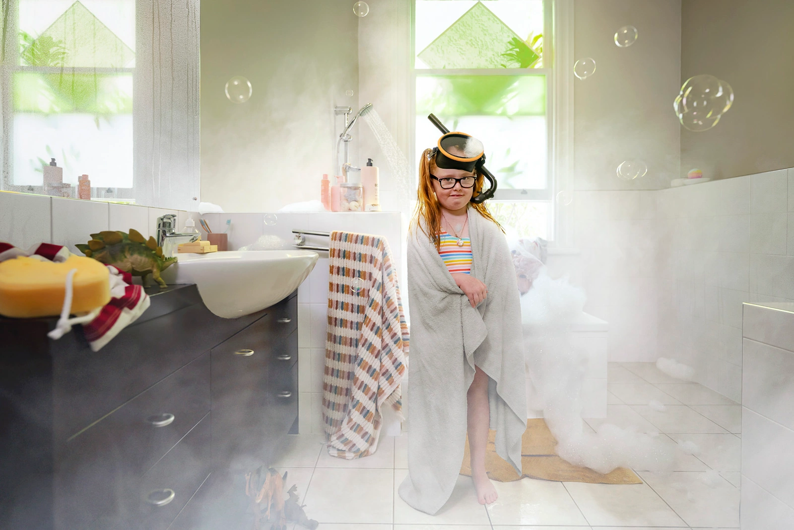 Young child wrapped in a grey towel wearing a snorkel on their head inside a steamy bathroom with soap bubbles floating.