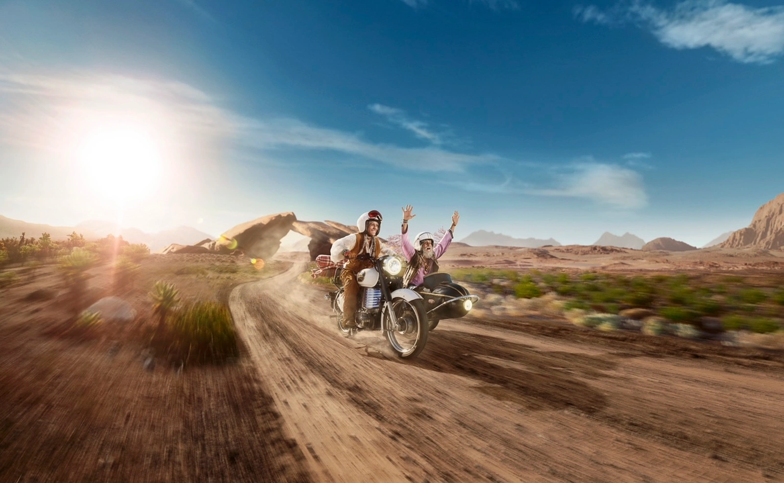 Two riders in white helmets on a vintage motorcycle with sidecar driving along a dusty dirt road in a desert landscape