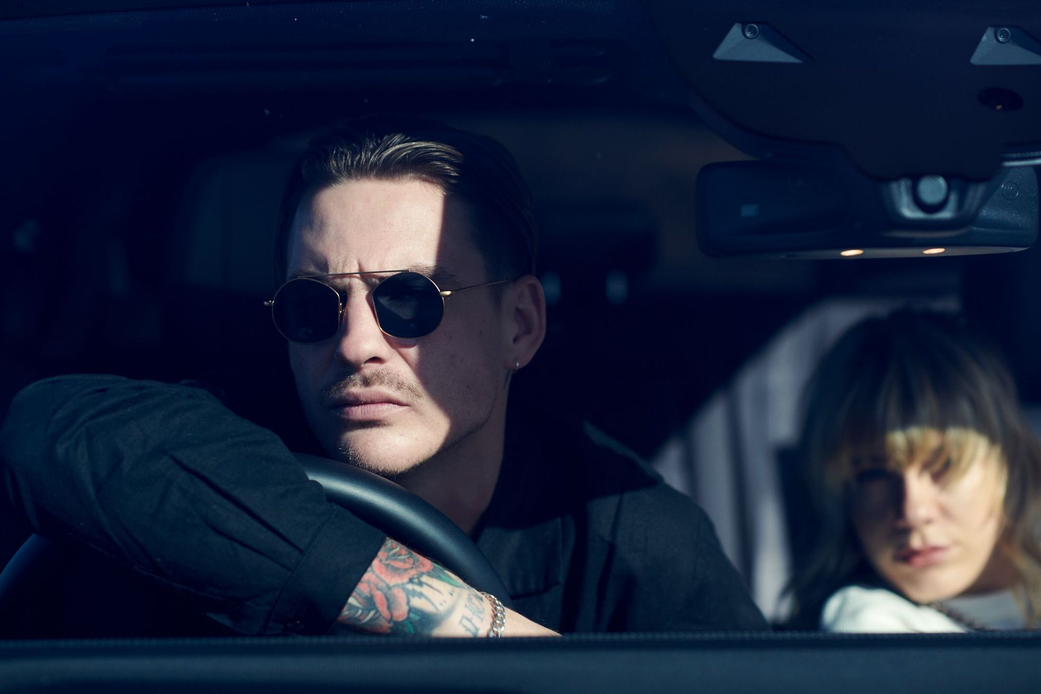 Man with tattoos and sunglasses driving car next to woman. Automotive setting with focus on driver and passenger in daylight.