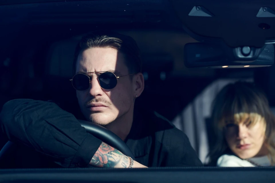Man with tattoos and sunglasses driving car next to woman. Automotive setting with focus on driver and passenger in daylight.