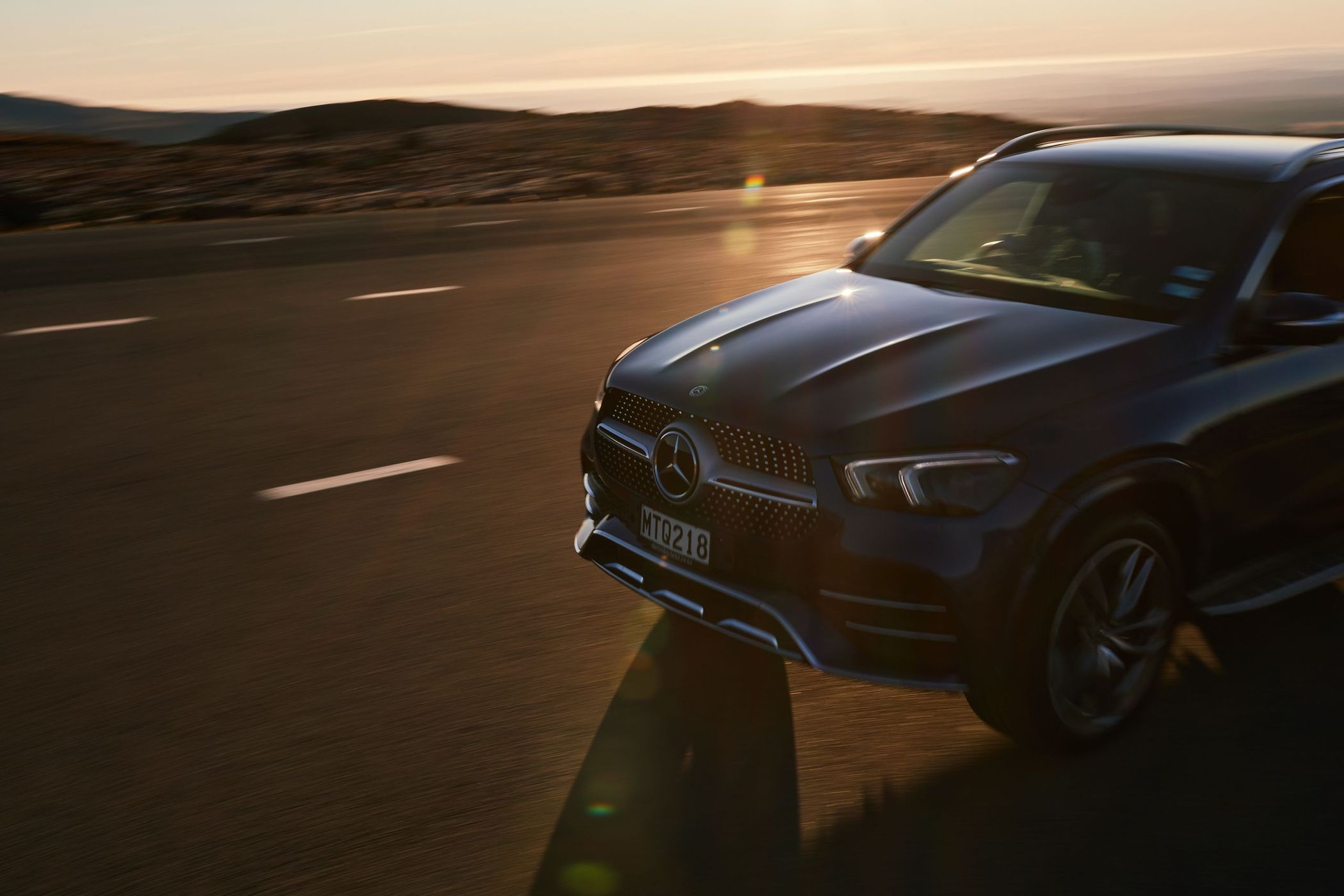 A dark blue Mercedes SUV cruises down an open highway at sunset, featuring mountains in the backdrop, automotive, and golden hour lighting.