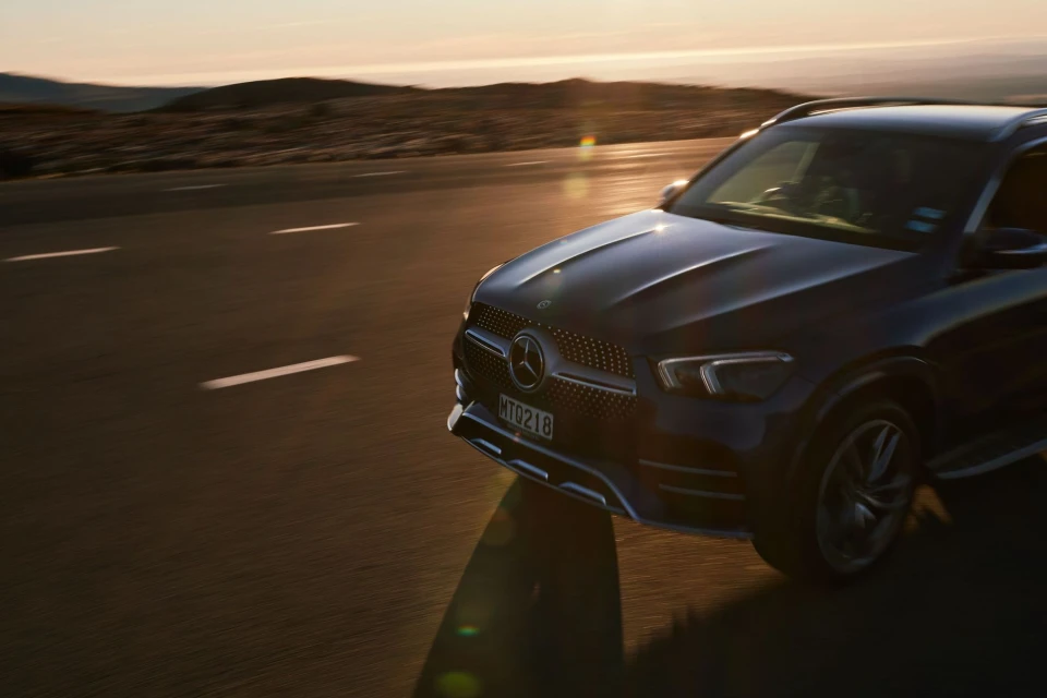 A dark blue Mercedes SUV cruises down an open highway at sunset, featuring mountains in the backdrop, automotive, and golden hour lighting.