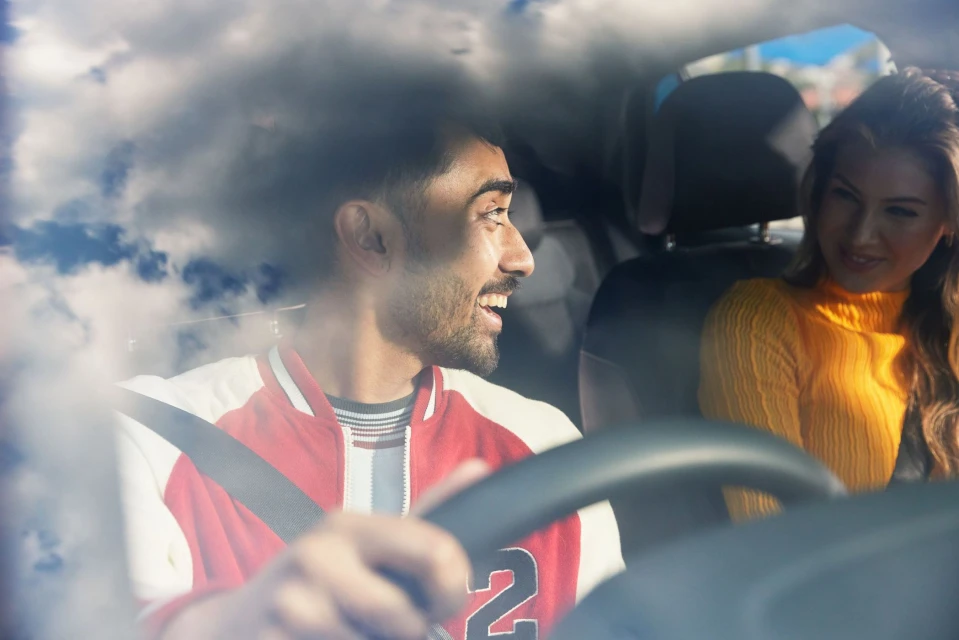 A smiling man drives a car with a woman passenger, both wearing seatbelts, in daytime with a cloudy sky seen through the windshield.