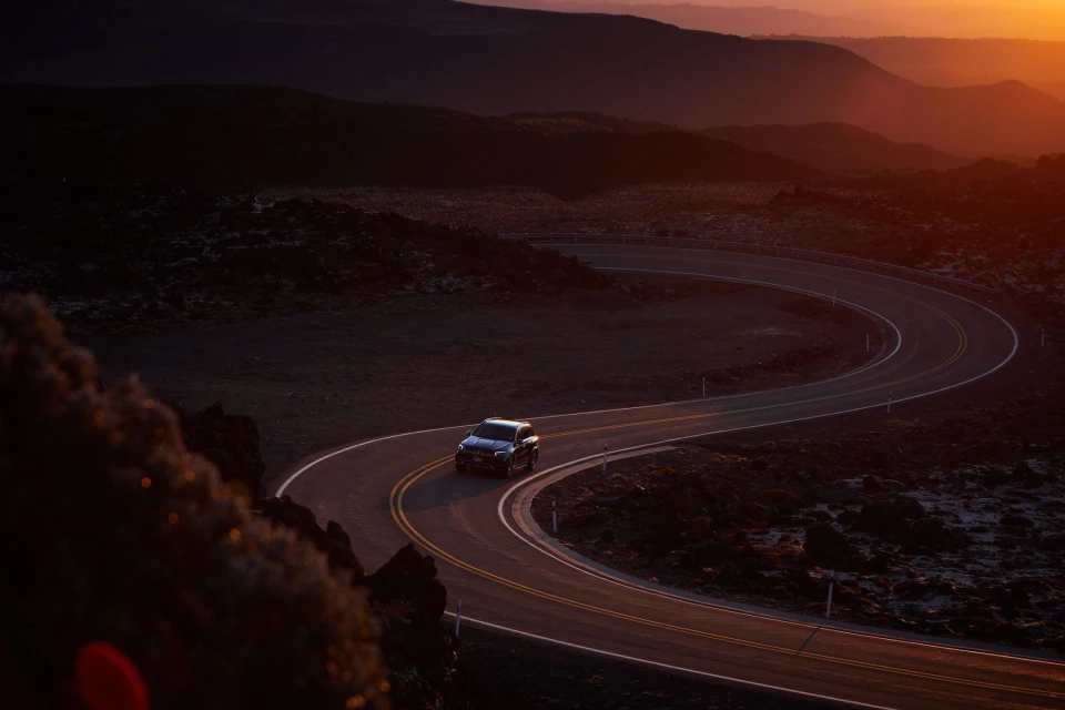 SUV driving on winding asphalt road through a dark, rocky landscape with mountains and sunset in the background. Car on scenic road.