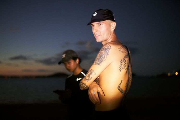 Man with tattoos wearing cap stands near beach with blurred figure in the background, illuminated by evening light.