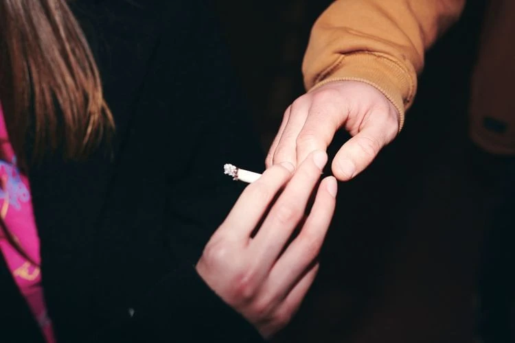 Close-up shot captures two people's hands in a dark environment, one offering a lit cigarette to the other, featuring a mustard jacket sleeve.