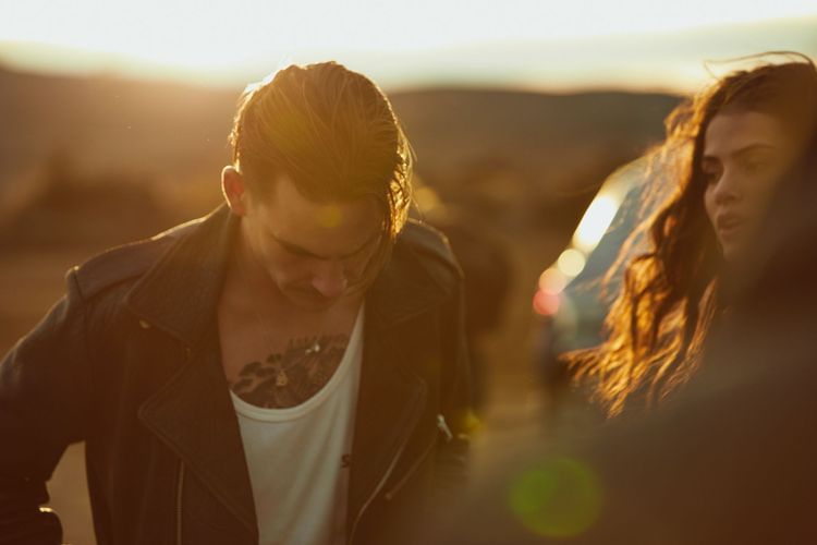 Outdoor fashion shot of a tattooed man in a leather jacket with a woman, bathed in warm sunset light in a desert landscape.