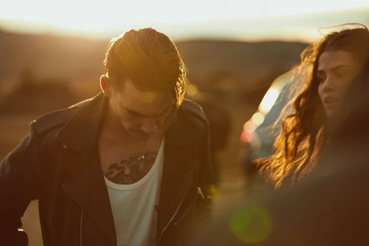 Outdoor fashion shot of a tattooed man in a leather jacket with a woman, bathed in warm sunset light in a desert landscape.
