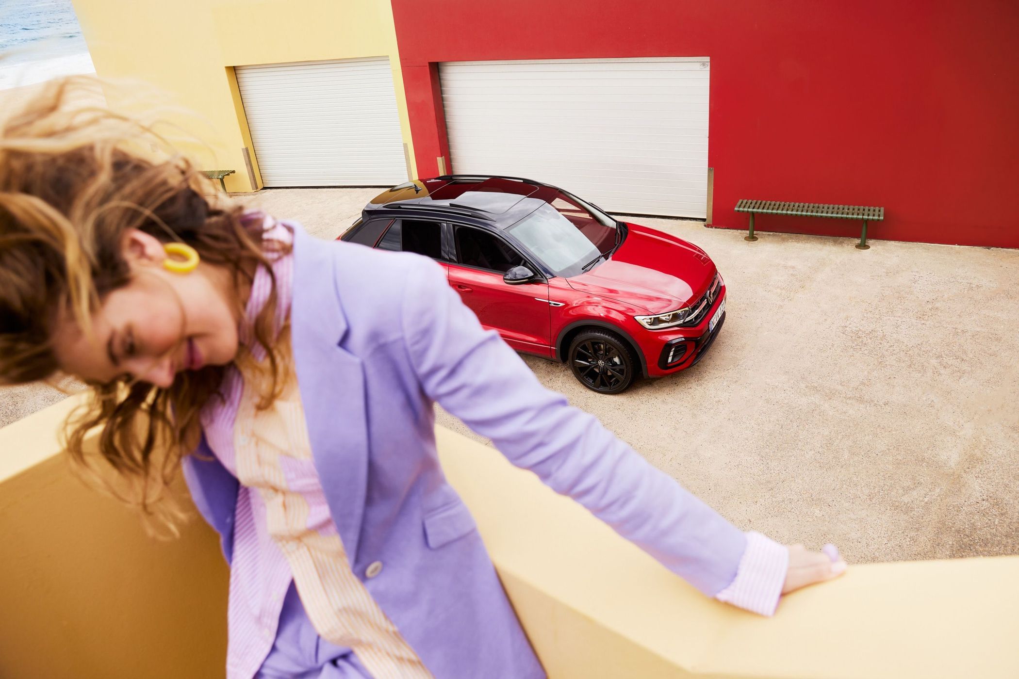 Stylish adult woman in purple suit poses near a red compact SUV with a black roof. The car is parked near colorful buildings.
